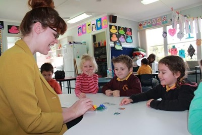 Teacher sits at desk with three young children doing activities with coloured paper