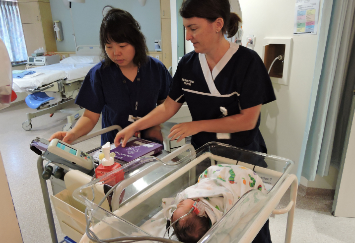 Two midwives check a newborn baby's hearing in a hospital crib