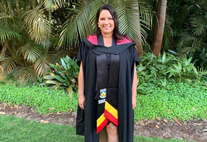 A young woman stands in front of tropical plants wearing a University graduation gown and smiling.