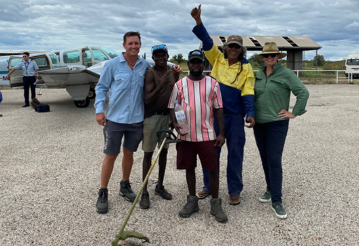 a group of five people stand on a regional airstrip, one in high viz has a thumbs up in the air. Another hods a whipper snipper and there is a small plane in the background. 