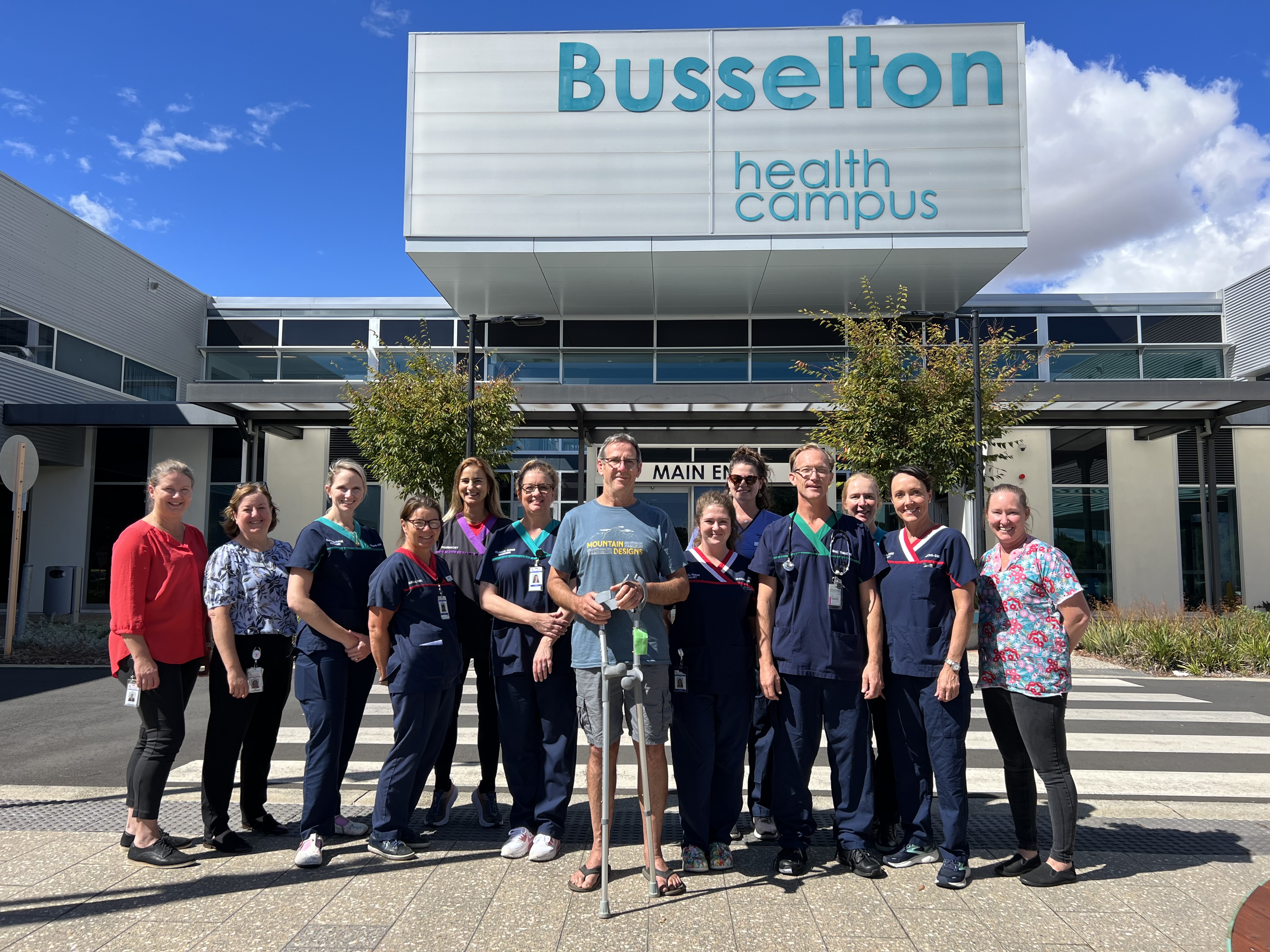 Knee replacement patient Peter stands out the front of Busselton Health Campus on a bright, sunny day surrounded by hospital staff.