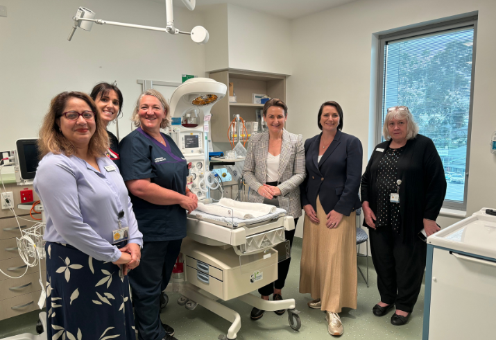 Six women stand in groups of three on either side of a PANDA cot for newborns.