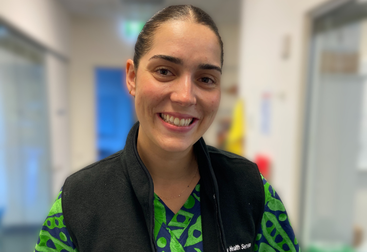 A dark haired woman smiles, stanidng in a busy hospital corridor