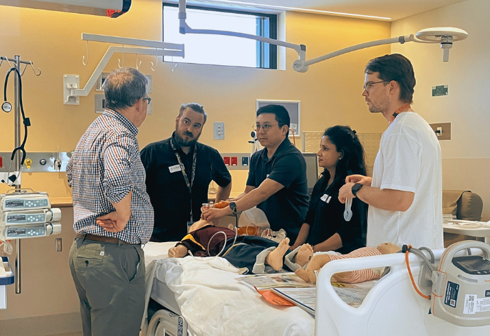 A group of medical professionals in discussion stand around a hospital bed with medical equipment 
