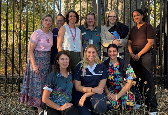 A group of nine women some in nurses scrubs are gathered by a fence under a boab tree, three of them are kneeling in front