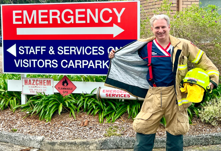 Man in fire fighter uniform with nursing scrubs underneath standing in front of hospital emergency sign.