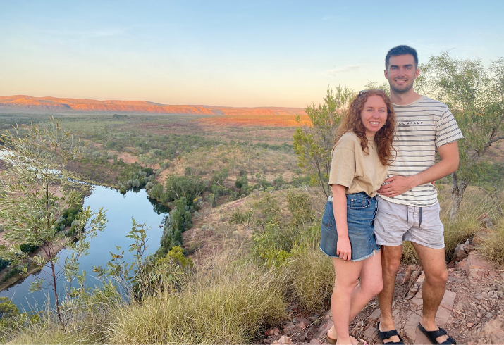 Man and woman standing on a hill with bush and a river behind them at sunset.