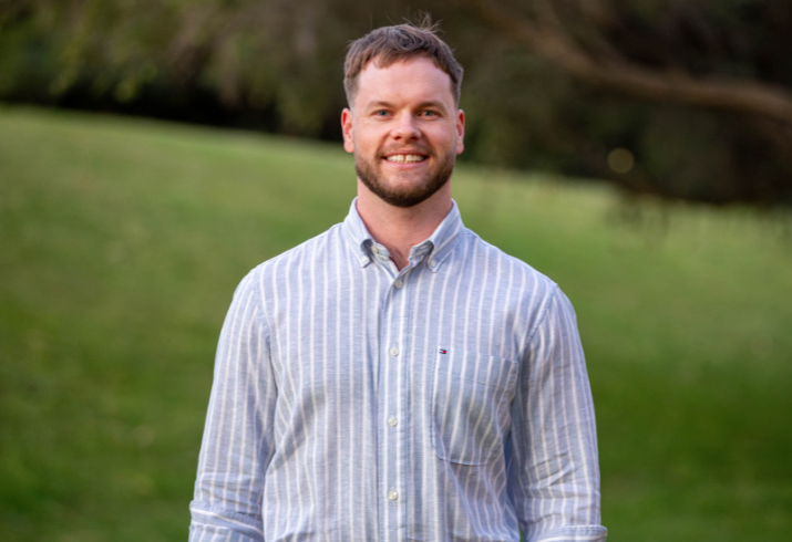 Man in striped button up shirt standing in front of green grass hill and tree.
