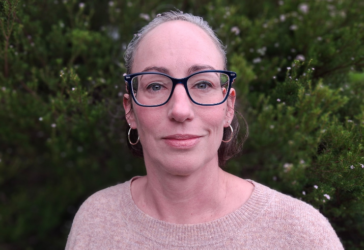 A bespectacled woman in a light pink top stands in front of a verdant green hedge