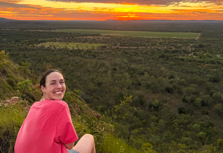 Woman in pink shirt on top of a large green hill looking out over the bushland and horizon.
