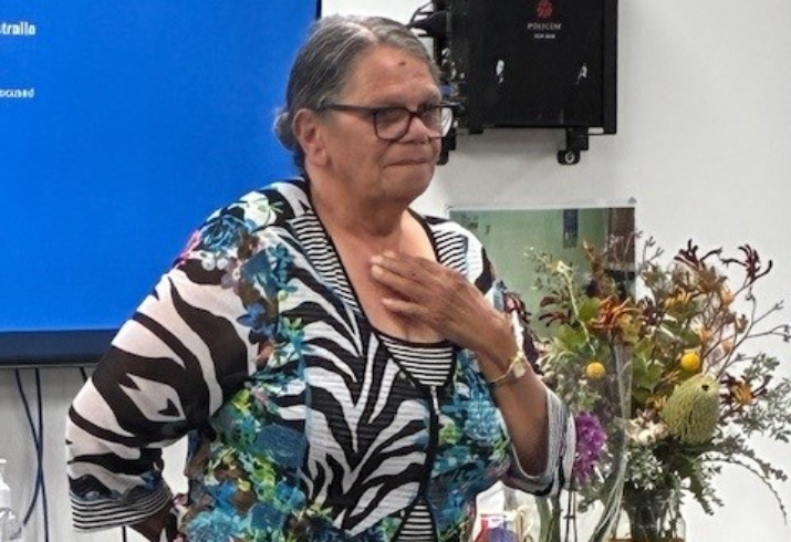 Woman standing in front of a presentation screen addressing the audience with hand on chest.