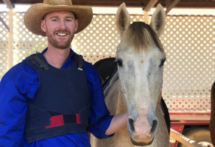 Man in blue shirt and akubra hat standing next to a white/cream horse.