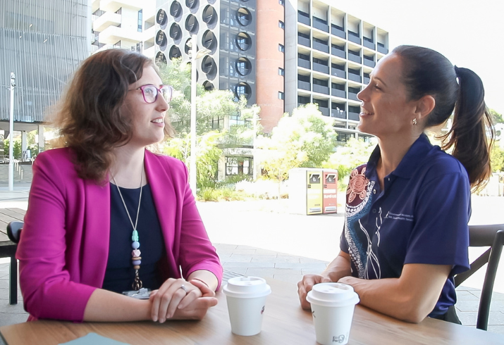 Two women sit opposite one another at an outside cafe with takeway coffees in front of them, they face each other in conversation.