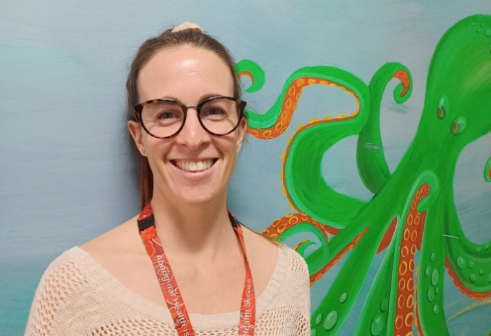 A smiling woman with glasses and hair pulled back stands against a painted wall with a bright green cartoon octopus on it
