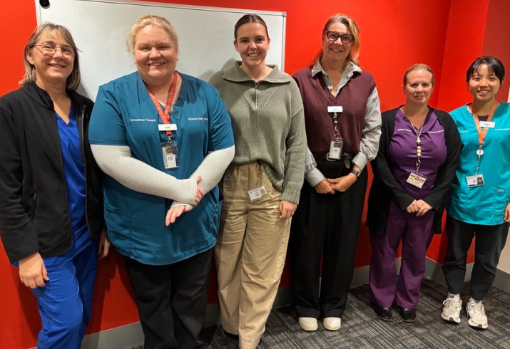A group of six nurses in various forms of attire stand against a red wall smiling 