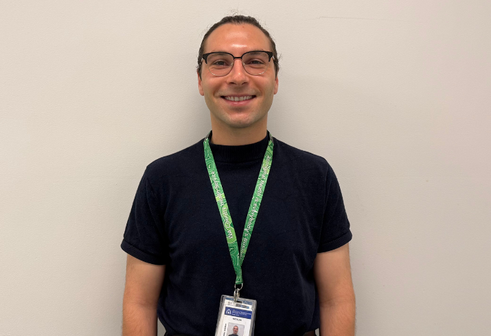 Doctor in a black shirt with a green lanyard and glasses against a white wall.