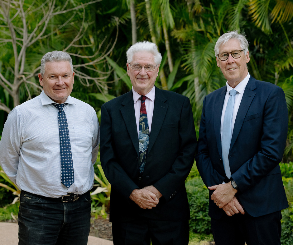 Three me stand together for a professional posed photo, smiling in business casual dress. They are outside, trees can be seen behind.