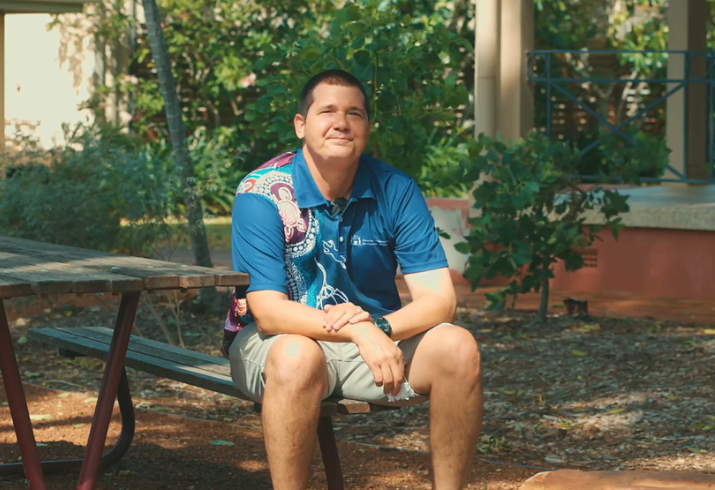 Man in polo shirt sitting on a bench outside.