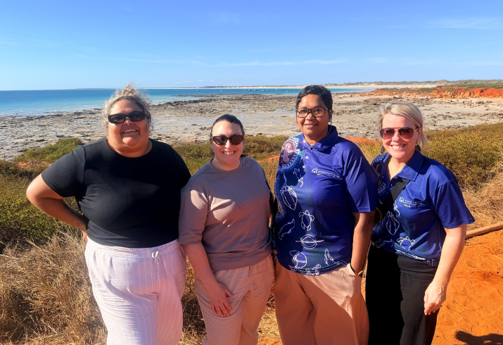 Four women stand overlooking the north west coast of Western Australia, it is a bright sunny day and the surrounding landscape is quite barren
