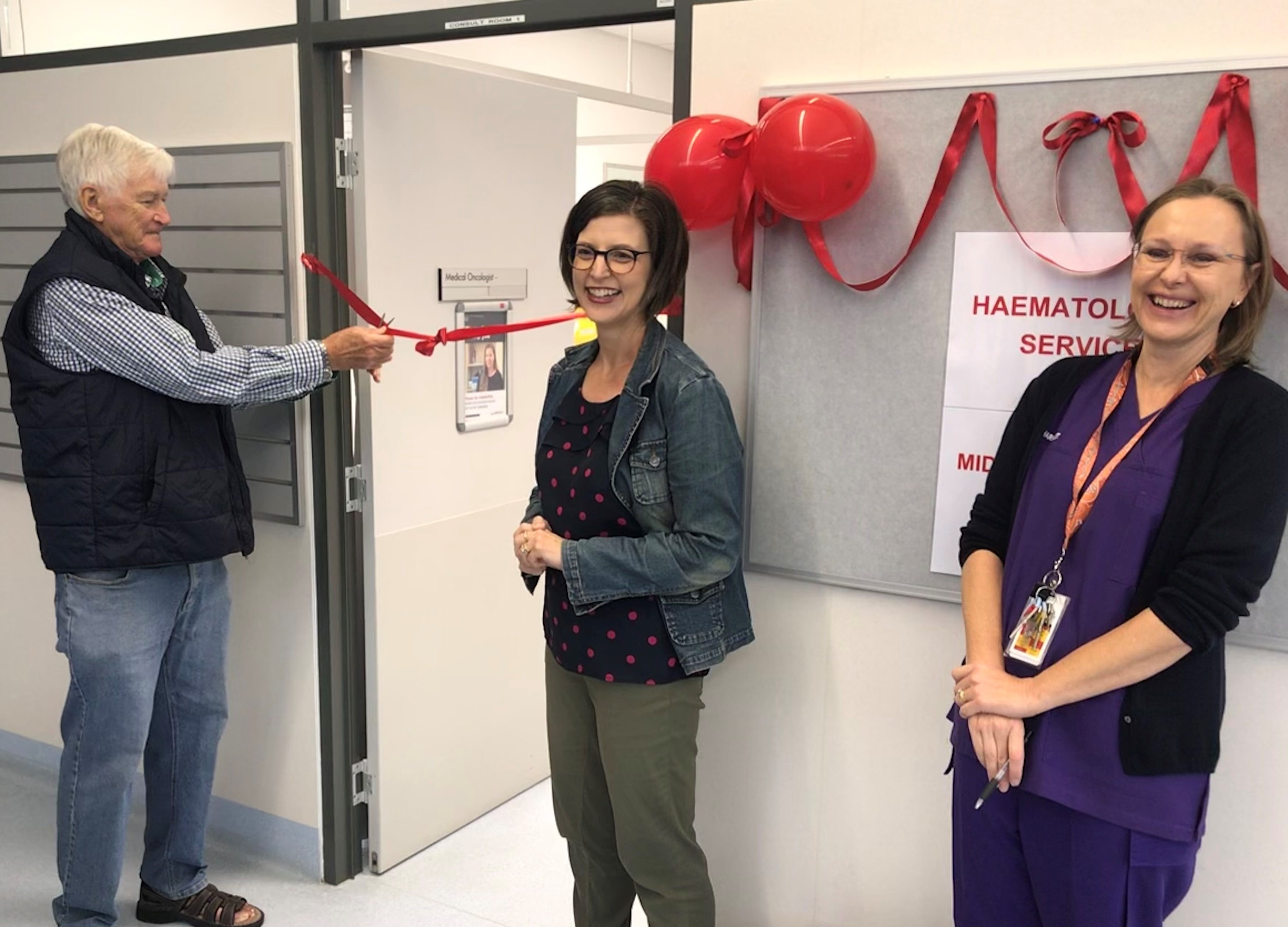 Patient John cuts the ribbon opening  Midwest Haematology with Dr Rebecca Howman and Clinical Pharmacist Nicky Goslin smiling and looking on.